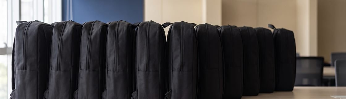 Fileira de mochilas MC115P idênticas alinhadas ombro a ombro sobre bancada clara, fugindo para o fundo desfocado