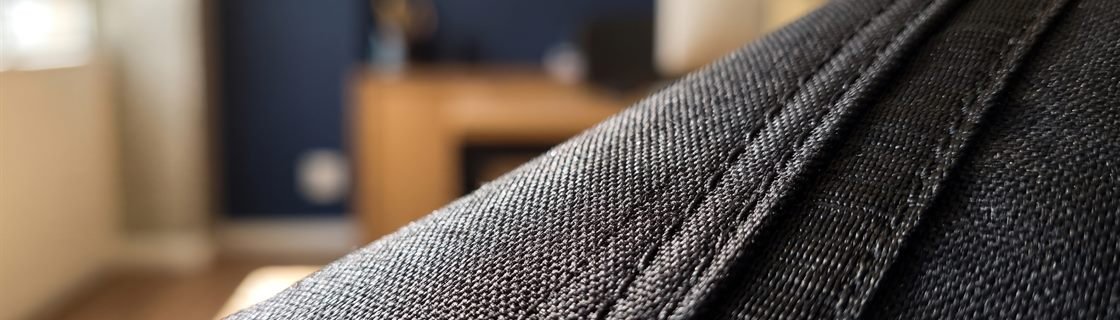 Macro da trama do poliéster da mochila MC115P em luz rasante, com os fios do tecido, a costura reforçada e a fita de reforço atravessando o quadro