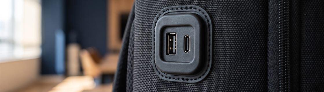 Close-up das entradas USB-A e Tipo-C embutidas na lateral da mochila MC700P, com o alojamento emborrachado e a trama do poliéster 900D ao redor